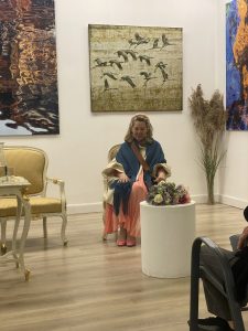 Ingrid Plater-Syberg siedzi na fotelu w galerii podczas wernisażu, w tle jej prace fotograficzne przedstawiające ptaki i wodę