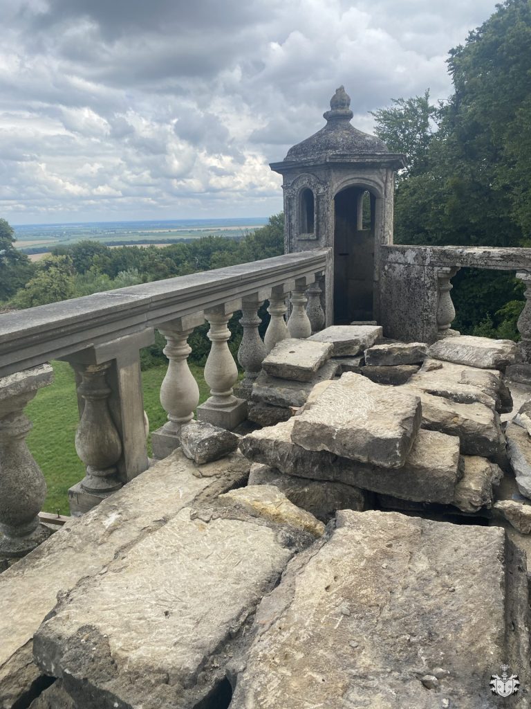 Widok z tarasu na koronie bastionu zamku w Podhorcach – demontowana kamienna nawierzchnia, kamienna balustrada z tralkami i wieżyczka narożna (gloriette) w tle, za nią rozległy krajobraz pól.