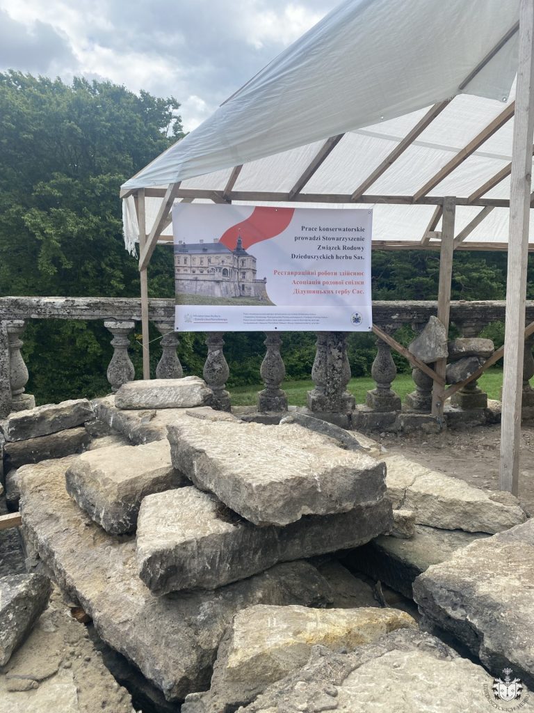 Baner informacyjny pod prowizorycznym zadaszeniem na tarasie zamku, z napisem po polsku i ukraińsku o pracach konserwatorskich prowadzonych przez Związek Rodowy Dzieduszyckich herbu Sas; w tle zdemontowane płyty kamienne i balustrada.