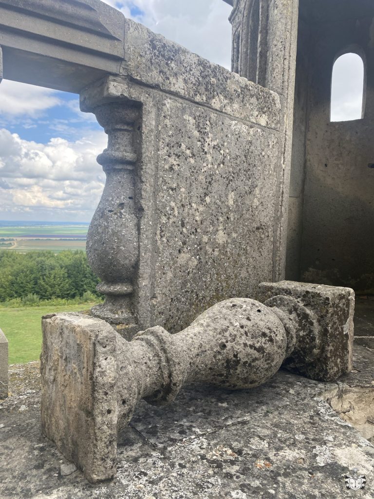 Zbliżenie na narożnik kamiennej balustrady tarasu – stojąca tralka oraz leżąca obok, zdemontowana tralka złożona z fragmentów; w tle otwór okienny w murze i widok na pola.