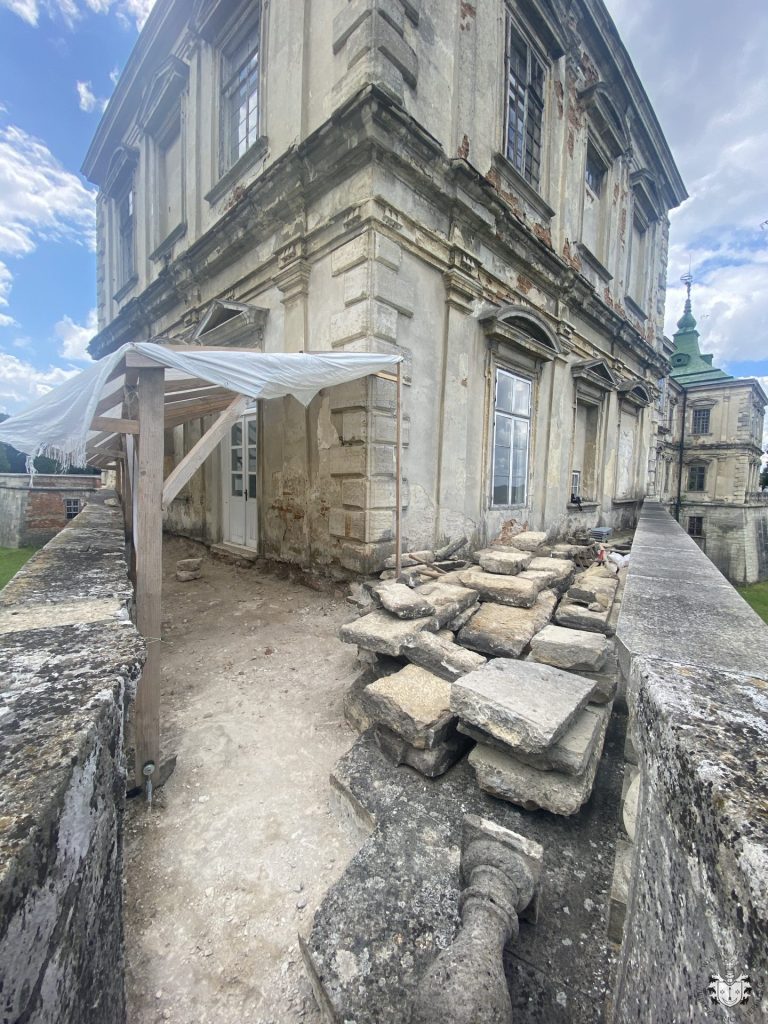 Narożnik elewacji zamku w Podhorcach ze zniszczonym tynkiem i odsłoniętą cegłą, prowizorycznym zadaszeniem nad wejściem, zdemontowanymi płytami tarasu ułożonymi na krawędzi muru.